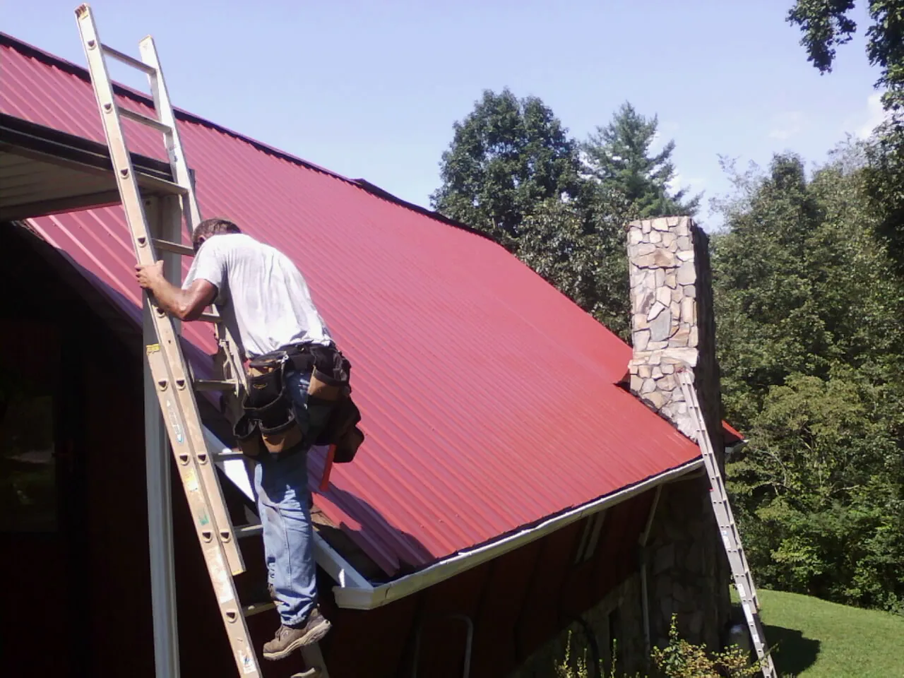 Roofer on a ladder beside a red metal roof and stone chimney — Soffit & Fascia Repair by Roofing Brothers in Orofino